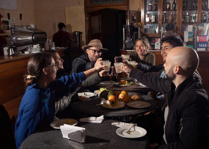 Six people sit around a table in a dimly lit restaurant, toasting with drinks. Plates of food and whole oranges are on the table. Shelves with bottles and a coffee machine are in the background.