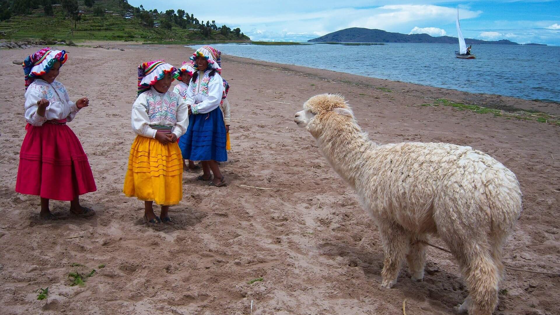Traditionally dressed girls from Llachón at the shores of Lake Titicaca with an alpaca and a sailboat. Visit Lake Titicaca in a sustainable fashion and get off the beaten path with RESPONSible Travel Peru.