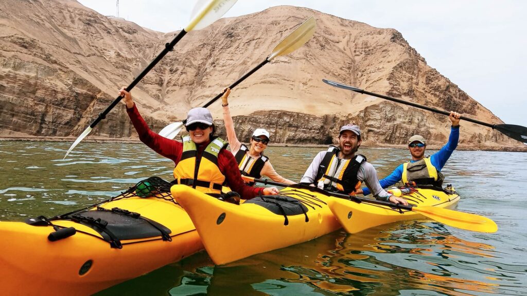 four kayakers in three kayaks saluting with paddles up in Paracas bay where the sea meets the the desert | Responsible Travel Peru