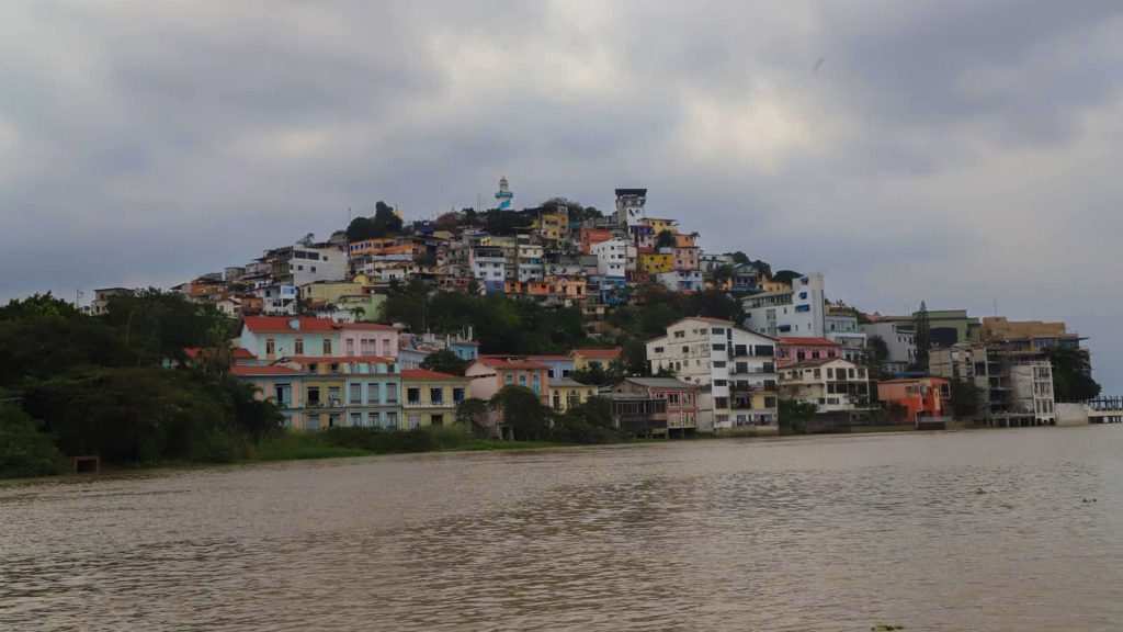 A hillside with colorful houses densely packed, set against a cloudy sky. A river flows in the foreground, bordered by green vegetation.