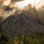 A volcano surrounded by dense vegetation beneath a dramatic sky with sun rays breaking through the clouds.