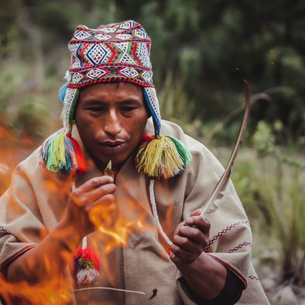 Person wearing a colorful hat and poncho sits by a fire, holding a stick and leaves, surrounded by blurred greenery.