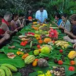 A group of people seated around a long table covered with tropical fruits and leaves, in an outdoor setting, engaging in a meal or ritual.