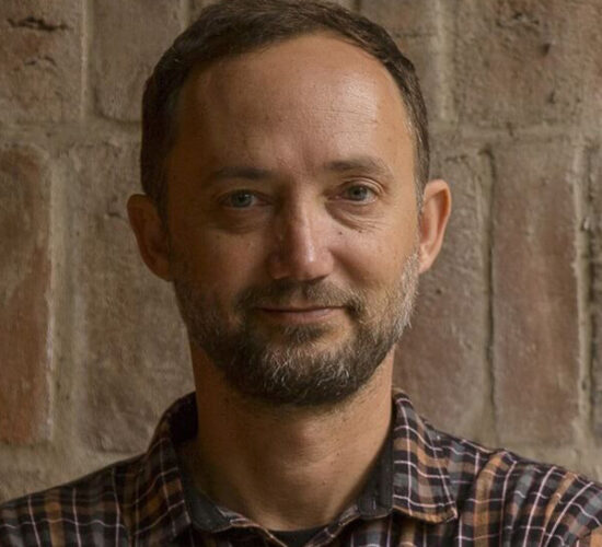 Man with short hair and a beard wearing a plaid shirt stands in front of a brick wall, looking at the camera.