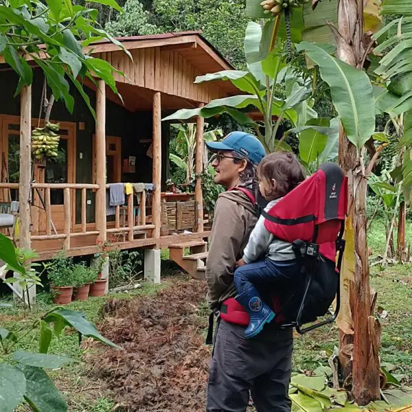 An adult wearing a cap carries a child in a red backpack carrier while standing in a lush garden near a wooden cabin surrounded by green plants and banana trees.
