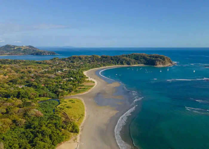 Aerial view of a curved sandy beach, lush forest, and blue ocean under a sunny sky—authentic Impactful Travel for sustainable tourism.